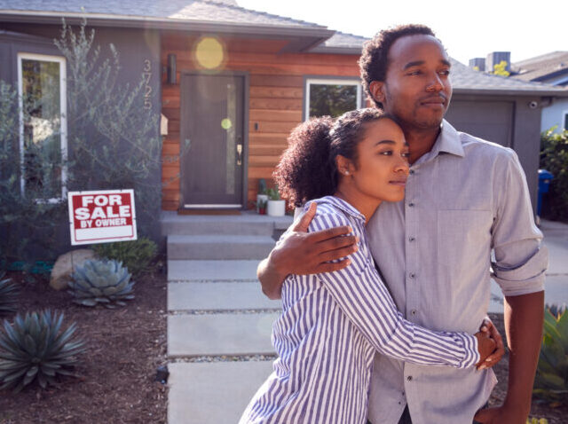 Thoughtful Couple Standing Outdoors In Front Of House With For Sale Sign In Garden