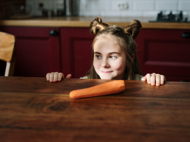 A girl smiles as she stands at a table with a carrot on it.