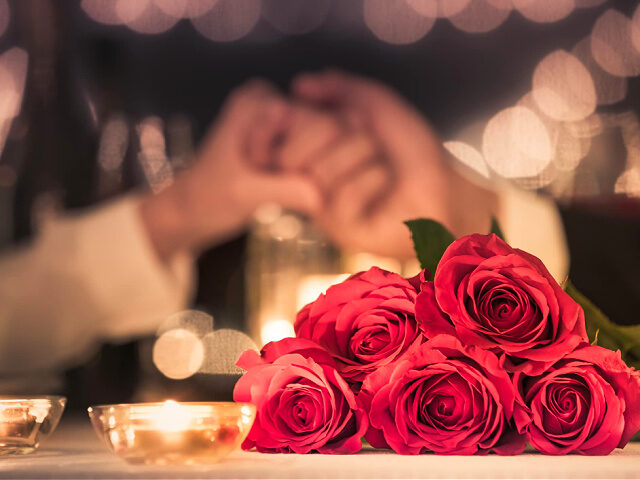 Couple at a candle light dinner date holding hands next to bouquet of red roses.