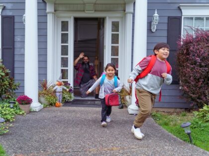 Elementary age Native American brother and sister run to meet the school bus as their dad waves goodbye from the front door of their home. (Fly View Productions via Getty)