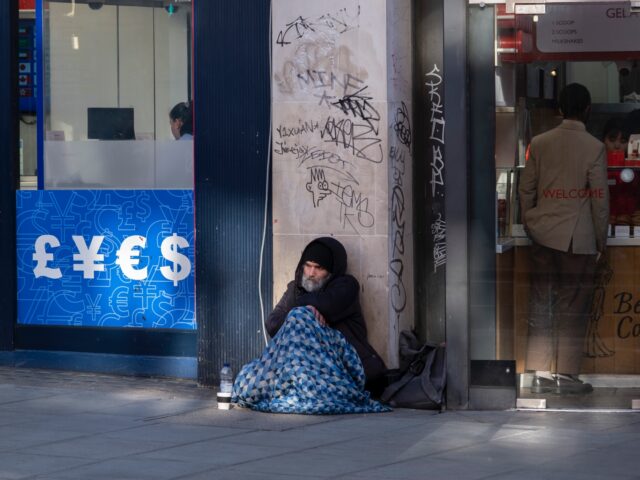 Man sits on the pavement under a sleeping bag beside a money exchange currency shop while