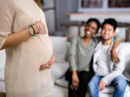 Pregnant Woman Touching Her Belly's Woman Standing In Front Of Smiling Young Couple Sitting