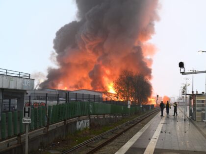 09 April 2023, Hamburg: Police officers stand at a stop at the Hamburg-Rothenburgsort train station while flames from a large fire can be seen in the background. Due to strong smoke and gas development, with possible chemical components, the local police have issued an official danger alert. Photo: Jonas Walzberg/dpa …