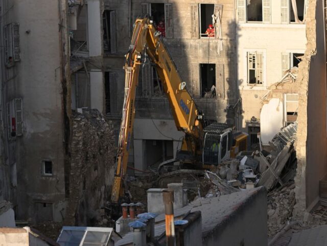 Firefighters look on from the windows of a nearby building as an excavator moves rubble at