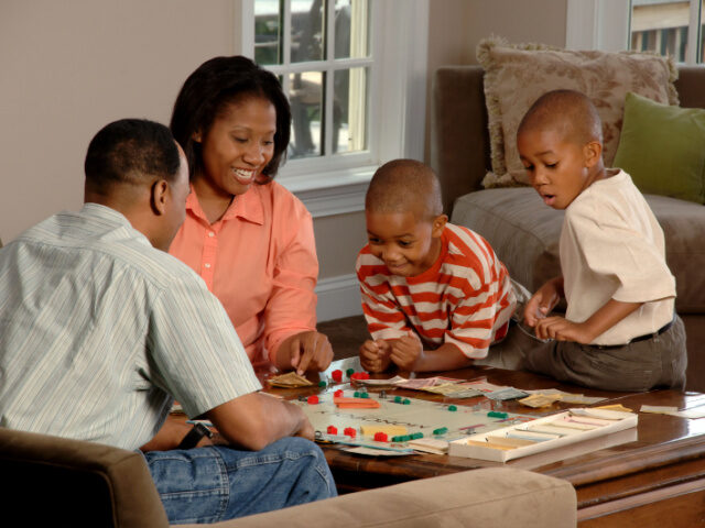 A family (husband, wife, and 2 children) sit around a coffee table playing a board game (U