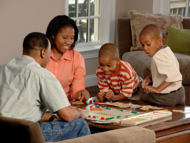 A family (husband, wife, and 2 children) sit around a coffee table playing a board game (Unsplash/ National Cancer Institute/Photographer Bill Branson)