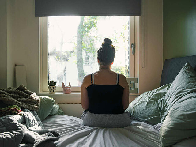 Rear view of a teenage girl looking out of the window whilst sitting on her bed - stock ph