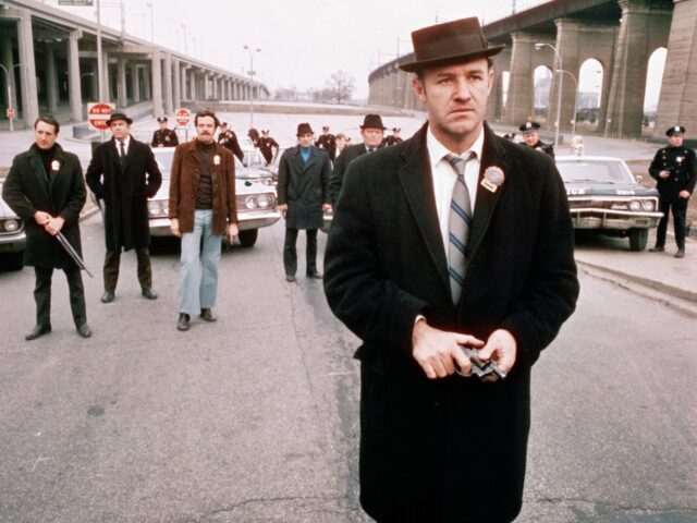 American actor Gene Hackman (foreground), as Detective Jimmy 'Popeye' Doyle, stands in the street by an overpass in front of a group of policemen and holds a gun in his hands in a still from the film 'The French Connection,' directed by William Friedkin, 1971. In the background is actor …