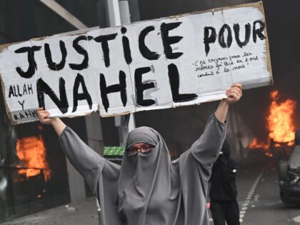 TOPSHOT - An attendee holds a banner reading "Justice for Nahel" as cars burn in the street at the end of a commemoration march for a teenage driver shot dead by a policeman, in the Parisian suburb of Nanterre, on June 29, 2023. Violent protests broke out in France in …