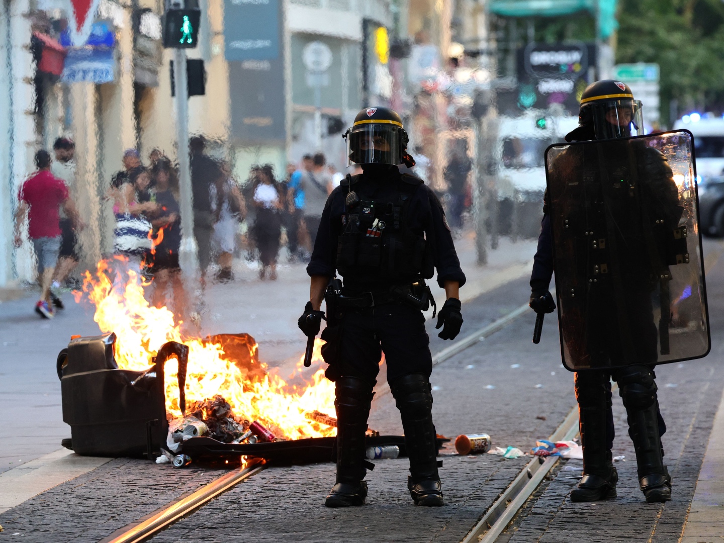 TOPSHOT - French riot police officers stand guard next to a burnt out trash bin during a demonstration against police in Marseille, southern France on July 1, 2023, after a fourth consecutive night of rioting in France over the killing of a teenager by police. French police arrested 1311 people nationwide during a fourth consecutive night of rioting over the killing of a teenager by police, the interior ministry said on July 1, 2023. France had deployed 45,000 officers overnight backed by light armoured vehicles and crack police units to quell the violence over the death of 17-year-old Nahel, killed during a traffic stop in a Paris suburb on June 27, 2023. (Photo by CLEMENT MAHOUDEAU / AFP) (Photo by CLEMENT MAHOUDEAU/AFP via Getty Images)