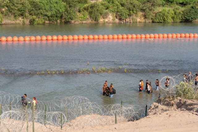 Migrants walk by a string of buoys along the Rio Grande border with Mexico in Eagle Pass,