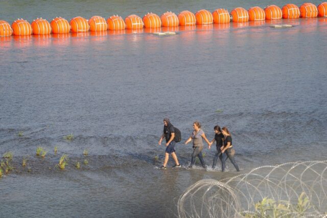 Migrants walk between razor wire and a string of buoys placed on the the Rio Grande border