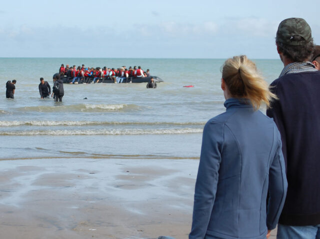 Members of the public watch as migrants sit onboard an inflatable boat before attempting t