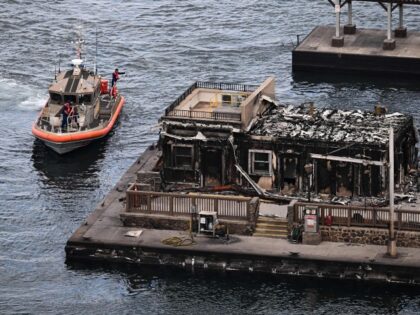 An aerial image shows a US Coast Guard vessel docking in the harbor near a destroyed build