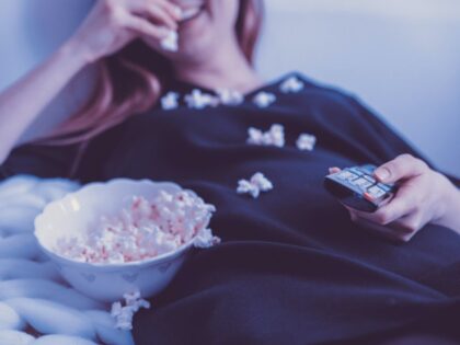 Smiling woman lies in bed watching TV and eating popcorn (Unsplash/jeshoots).