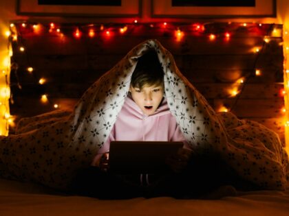 Boy lying on bed under blanket and using a tablet