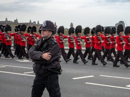 A policeman stands guard as soldiers from the British Army's Grenadier Guard bear their rifles as they march in full ceremonial uniform across Westminster Bridge to assume their positions for the elaborate ceremonies at the coronation of King Charles III in London, UK. The soldiers are wearing red tunics with …