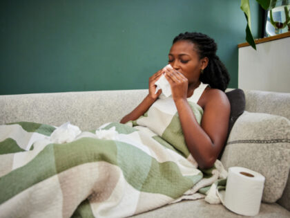 Young African woman with a cold sneezing into tissue while lying wrapped in a blanket on her living room sofa. Goodboy Picture Company via Getty Images.