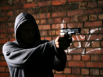 Someone adorned in a hoody points a gun as he stands beside a brick wall (Stock photo via