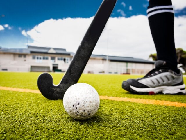 Hockey ball, hockey stick, pavilion, artificial turf. The ball is in sharp focus. Camera: