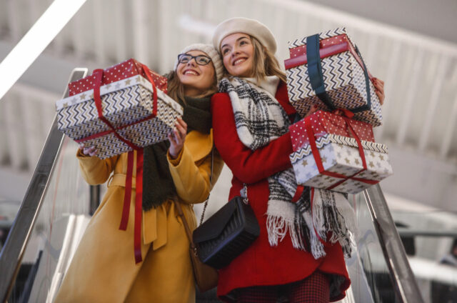 Two young women holding bunch of Christmas presents on descending escalator.