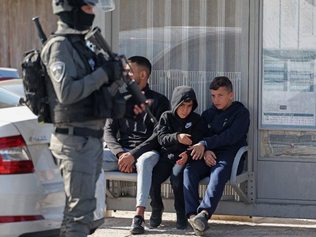 An Israeli border policeman stands guard next to children sitting on a bench at a bus stop