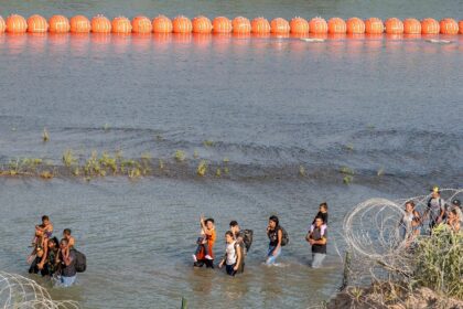 Migrants walk between concertina wire and a string of buoys placed on the water along the