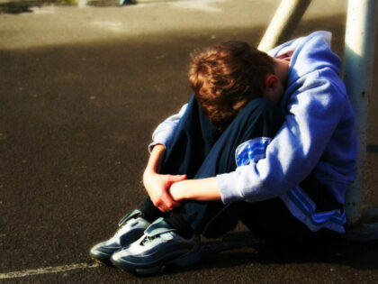 Boy head down hugging his knees on playground leant against goal post.