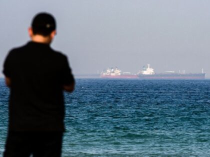 A man stands along a beach as tanker ships are seen in the waters of the Gulf of Oman off