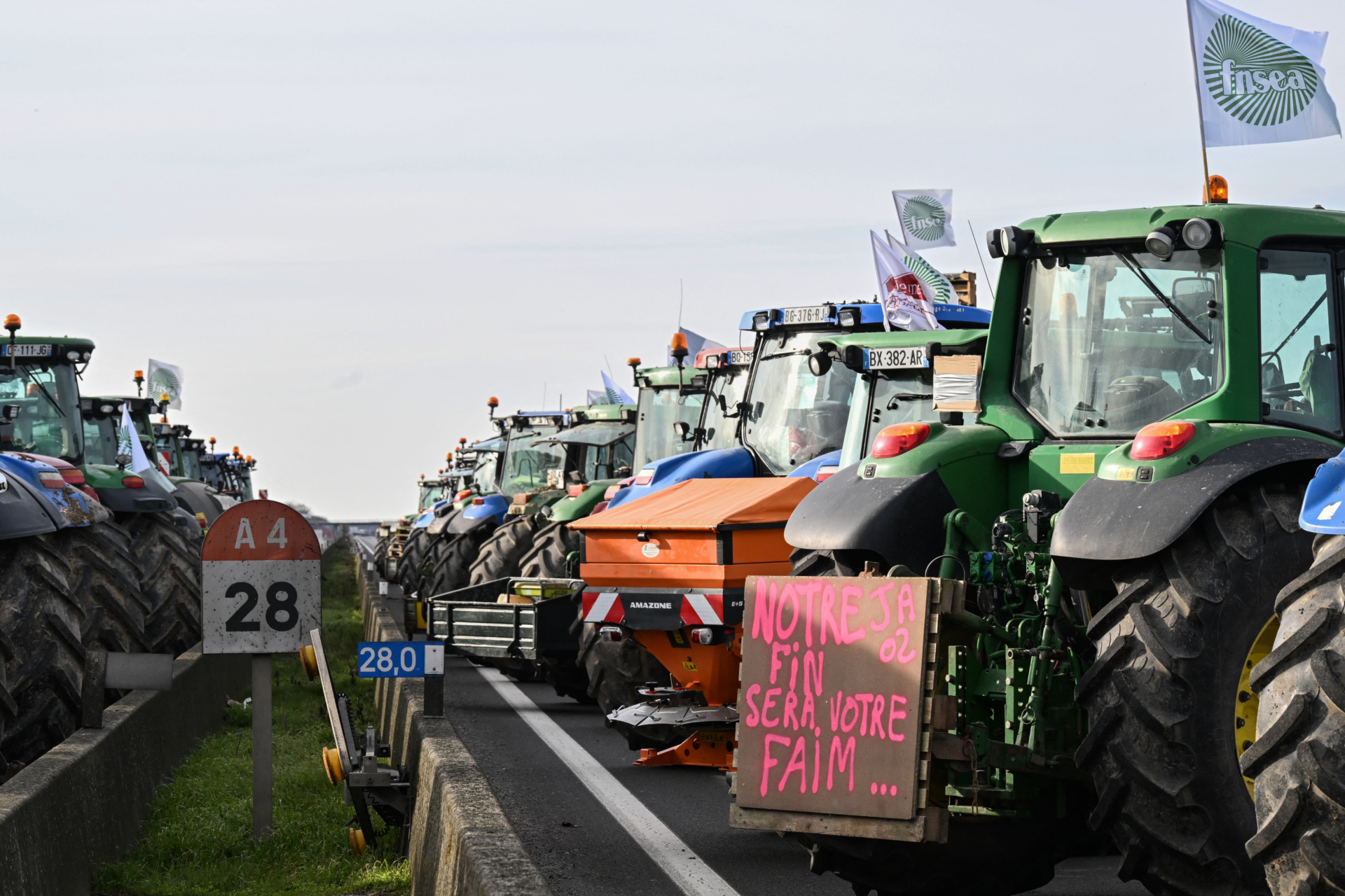 This photograph taken on January 29, 2024, shows tractors including one with a placard reading "our end will be your hunger" next to a road sign signaling the A4 highway, during a road block protest by French farmers on the A4 highway near Jossigny, east of Paris, amid nationwide protests called by several farmers unions on pay, tax and regulations. Local branches of major farmer unions FNSEA and Jeunes Agriculteurs announced on January 27, 2024, a "siege of the capital for an indefinite period" starting at 2 p.m. on January 29, 2024, as some farmers feel that the government's announcements in favor of the sector are insufficient. (Photo by Bertrand GUAY / AFP) (Photo by BERTRAND GUAY/AFP via Getty Images)