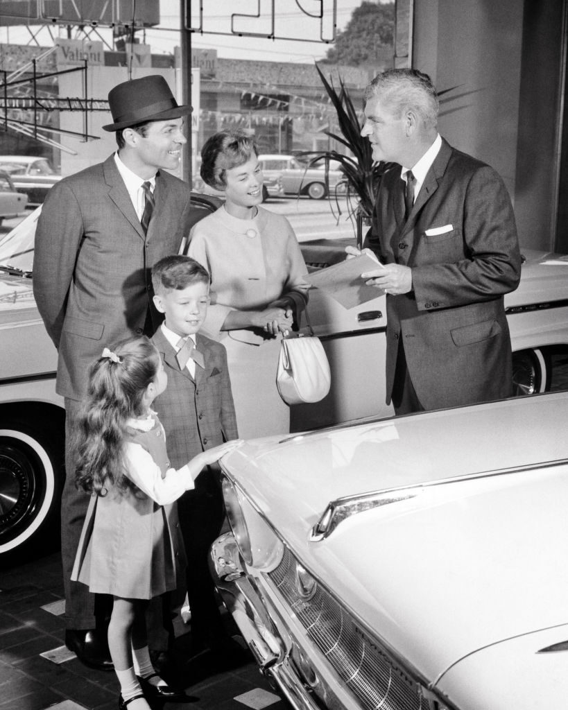 An American family in the 1960s look over the choices in a car showroom.1960s FAMILY DAD MOM BOY GIRL BUYING A NEW CAR WITH SMILING SALESMAN IN CAR DEALERSHIP SHOWROOM (Photo by H. Armstrong Roberts/ClassicStock/Getty Images)