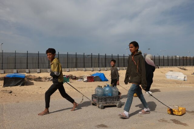 Palestinian children walk along a border fence as they carry water to a displacement camp