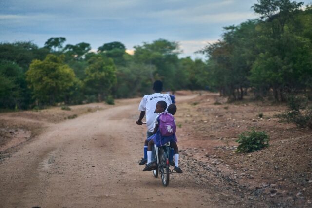 A relative pedals two students to school safely on a donated bicycle near Zimbabwe's Hwang