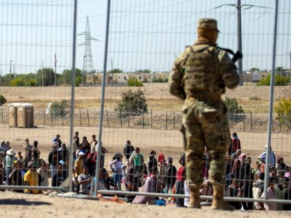 FILE - Migrants wait in line adjacent to the border fence under the watch of the Texas Nat