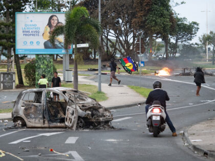 People walk next to a burnt-out car while a man (C) holds a flag of the Socialist Kanak Na