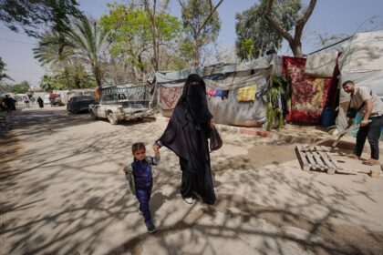 A displaced Palestinian woman holds a child by the hand as she walks in front of tents set