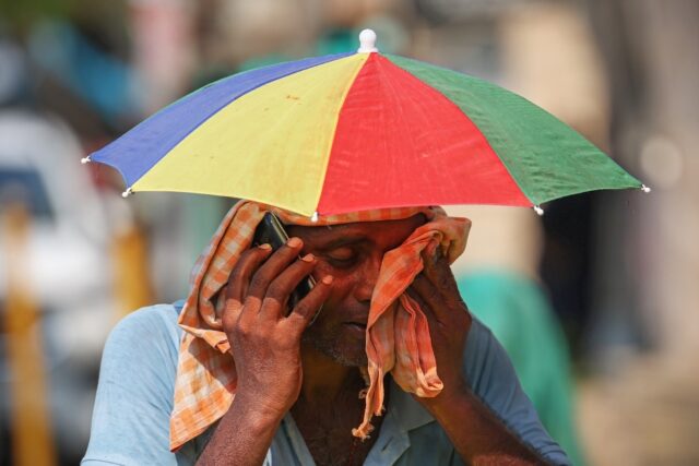 A vendor with an umbrella over his head talks on mobile phone as he wipes his face with a