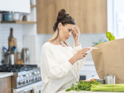 A young woman stands at the kitchen counter holding her grocery bill as she reviews it wit