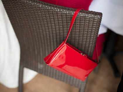 Red patent leather handbag on the back of a brown basket chair - stock photo