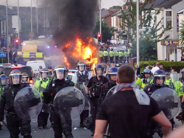 SOUTHPORT, ENGLAND - JULY 30: Riot police hold back protesters near a burning police vehic