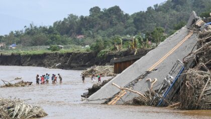 People cross a river next to a bridge that collapsed as the river surged due to rains brou