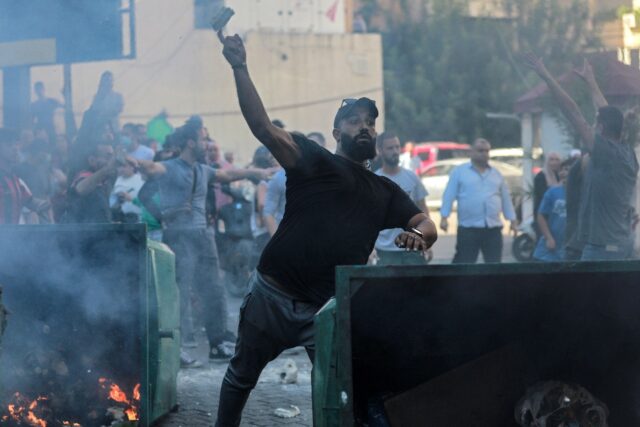 A man throws a stone from behind a burning rubbish container as displaced people who fled