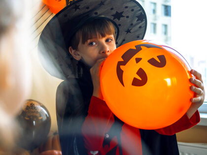 Boy in a black cape and a witch's hat blows a balloon with the image of a pumpkin jack.