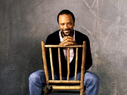 American record producer Quincy Jones smiles, sitting in a wooden chair with his cat in Lo