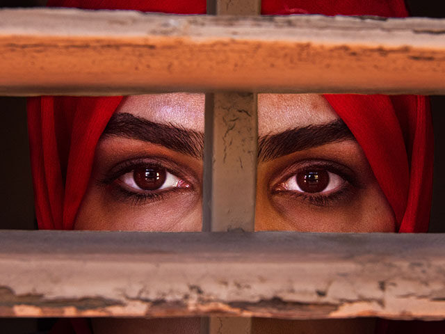 Liza Anvary, an Afghan refugee, poses behind a wooden window pretending to be in prison to