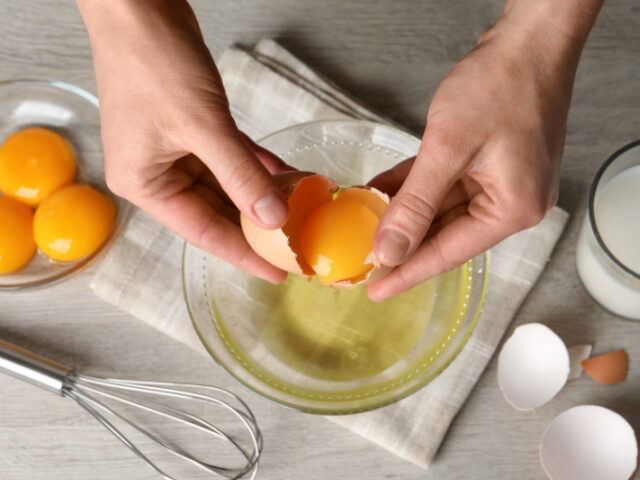 Woman separating egg yolk from white over glass bowl at light wooden table, top view - sto