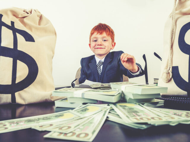 A young boy dressed as a businessman with a big pile of money. There are $100 bills and mo