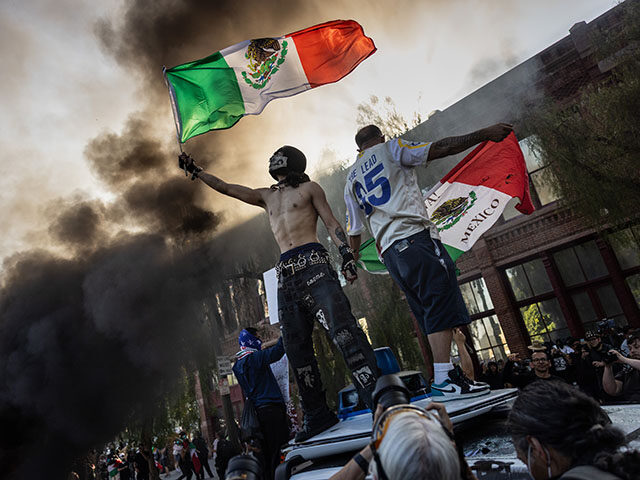 With smoke filling the air from several burning Waymo driverless cars, men are seen standi