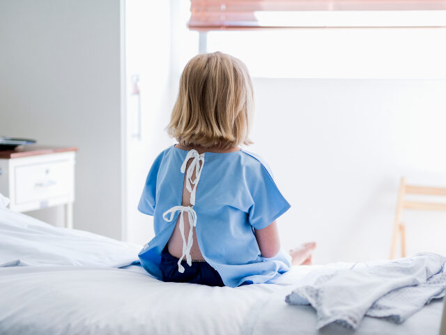 child in hospital Rear view of ill girl sitting on bed. Female child patient is wearing hospital gown. She i