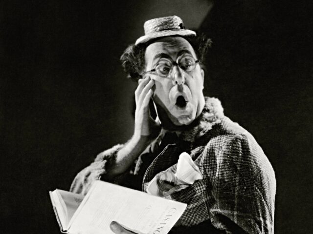 Comedian Ed Wynn, in a woman's plaid coat with a small straw hat and horn-rimmed glasses,